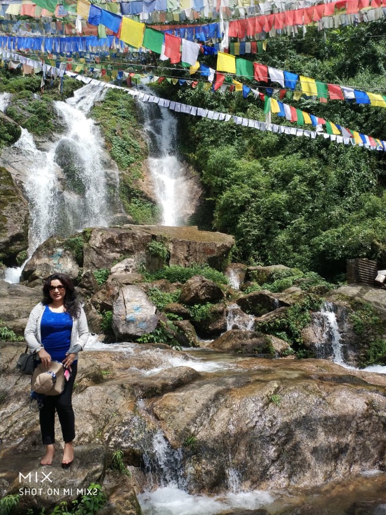Traveler beside Lhasa Falls surrounded by lush greenery and colorful Tibetan prayer flags