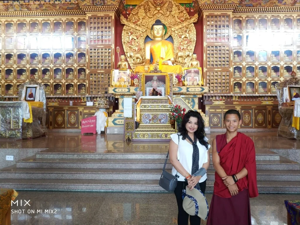 A woman standing next to a young Buddhist monk inside a stunning, ornately decorated monastery with a large golden Buddha statue